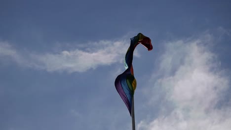 rainbow flag for lgbt gay community on pole and sky behind . is first droop but starts to wave swiftly. blue skies behind with some cloud. sunlight  makes flag's translucent fabric glow.
