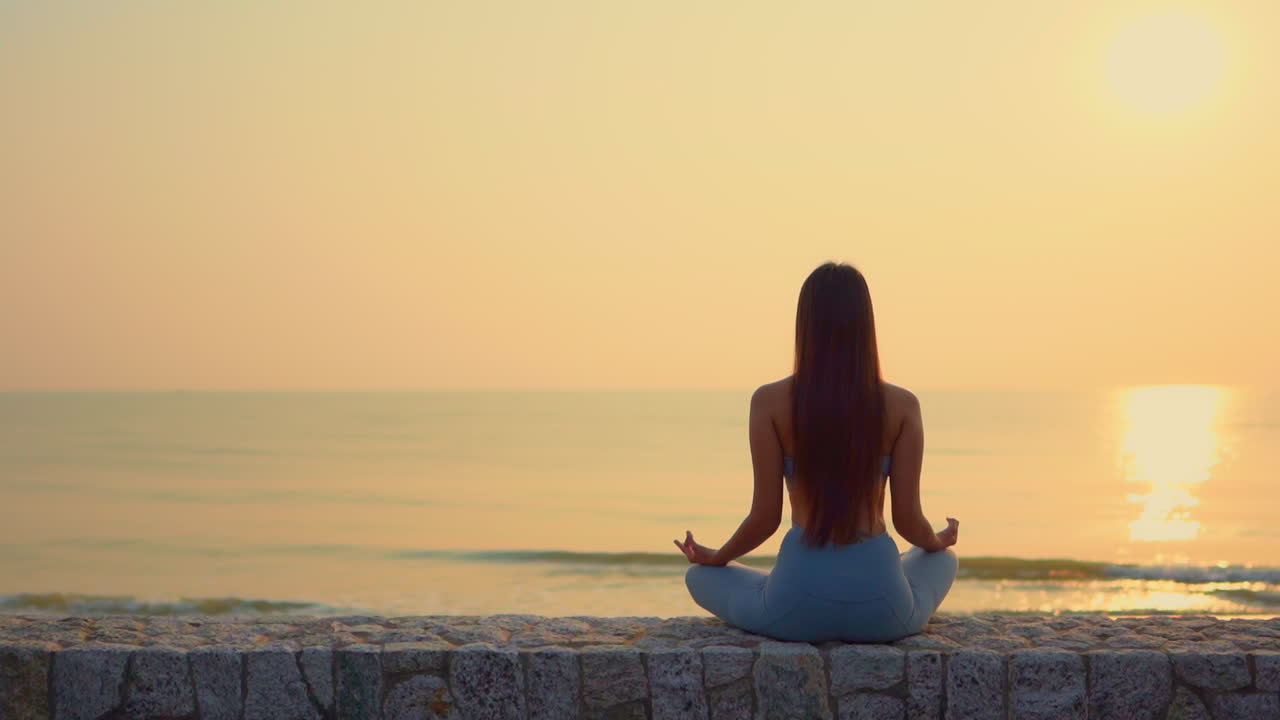 A woman sitting in the lotus position looks out over the ocean as the sunsets