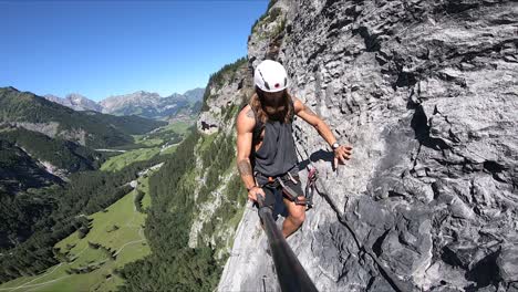 a tattooed man with beard and long hair is walking on the small, thin and dangerous edge of a mountain, moving his carabiner and safety rope along with him