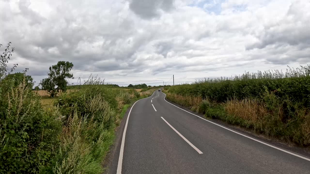 A vehicle travels down a narrow, winding rural road bordered by hedges and fields under overcast skies, with gentle camera movement and natural daylight