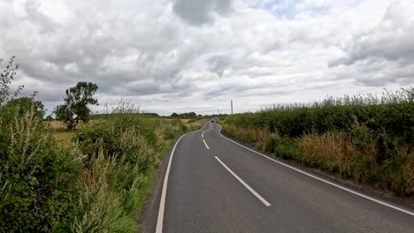 a vehicle travels down a narrow, winding rural road bordered by hedges and fields under overcast skies, with gentle camera movement and natural daylight