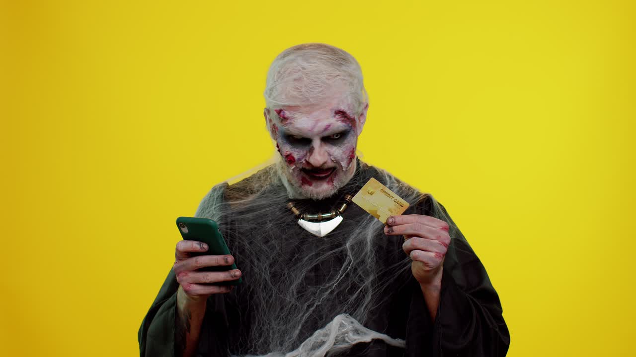 Uomo sinistro zombie di Halloween che usa carta di credito e smartphone durante gli acquisti online