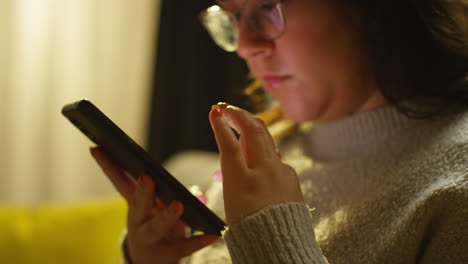 close up of young woman spending evening at home sitting on sofa with mobile phone scrolling through internet or social media 2