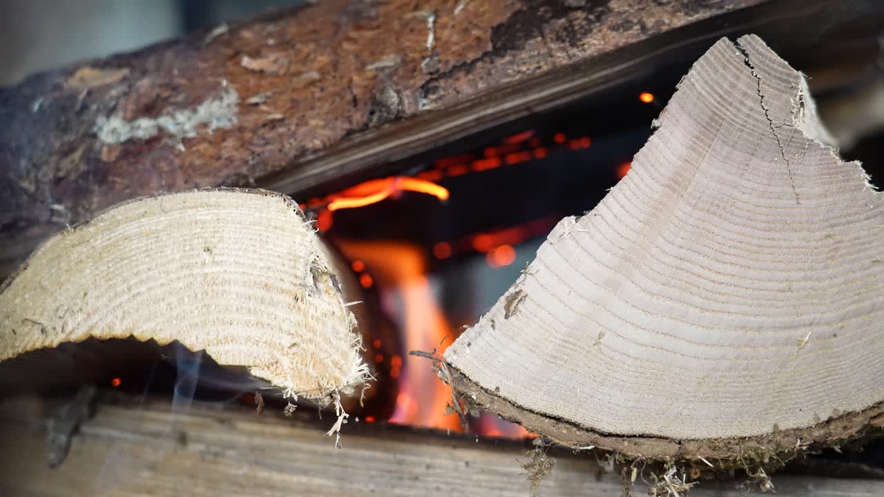 Close-up gerak lambat kayu bakar dengan serat kayu yang fokus tajam di latar depan, sementara nyala api berkedip lembut di latar belakang. Kontras tekstur dan cahaya api menciptakan suasana dalam ruangan yang hangat.