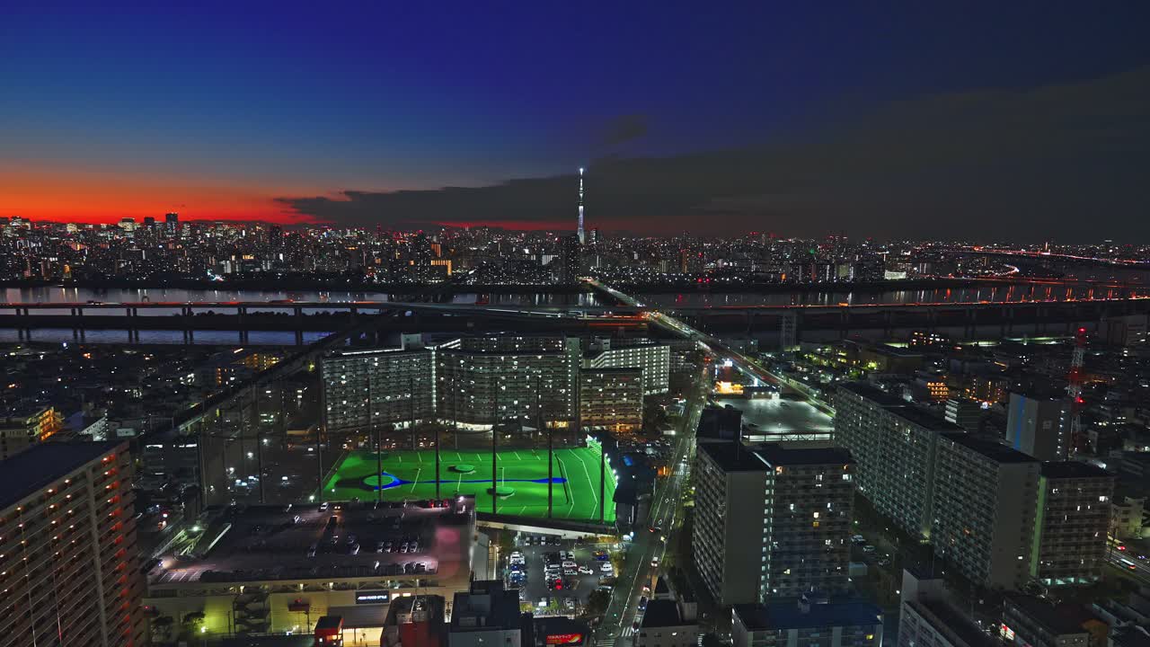 Karanlık bir yerleşim bölgesinin ortasında parlak yeşil ışıklı bir golf antrenman sahasını gösteren yüksek açılı bir çekim. Tokyo silüeti ve parlayan bir kule, canlı bir gün batımı gradyanının altında yükseliyor.