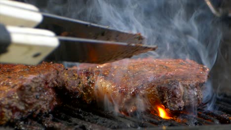 a pair of meat tongs slide a nearly cooked juicy rib eye steak on a grill and presses on it as flames shoot up in slow motion