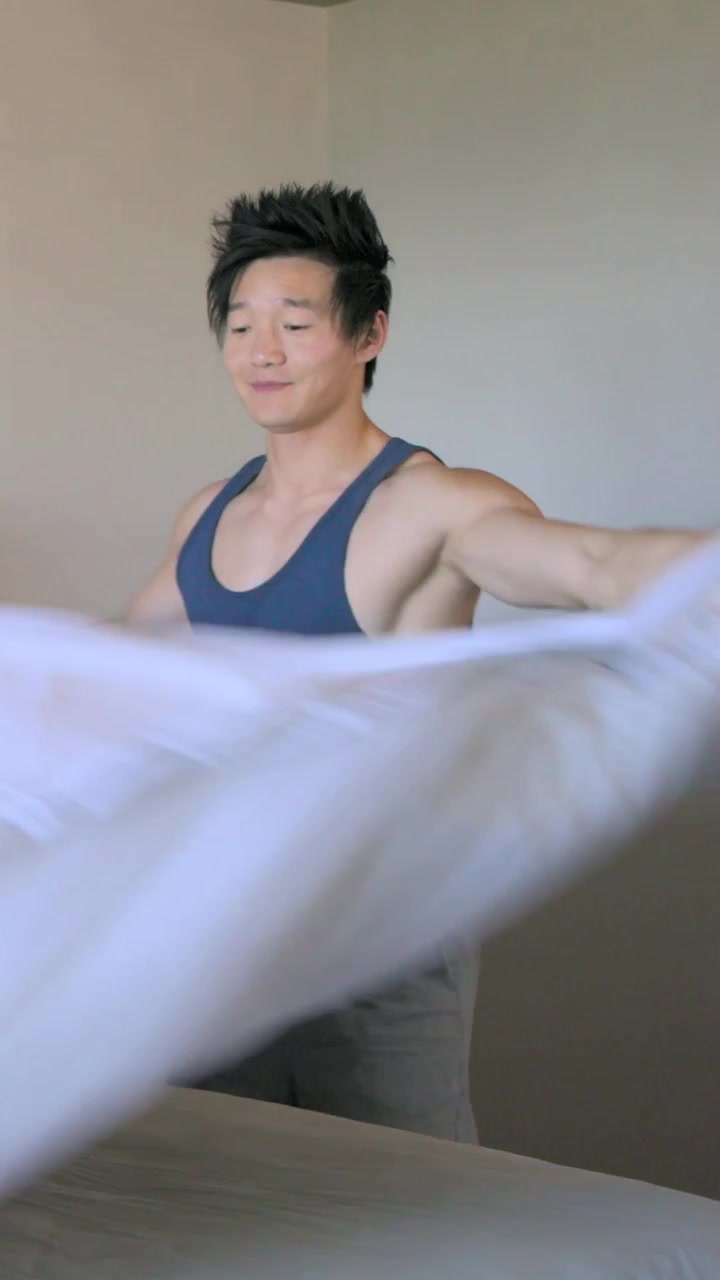 Video verticale: Uomo asiatico che si sveglia, si alza e rifà il piumone in camera da letto, indossando una canotta blu scuro.