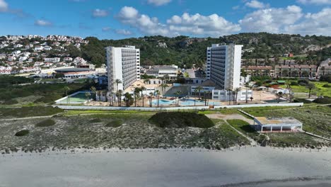 a cinematic aerial view of son bou beach with buildings in the background in menorca, spain