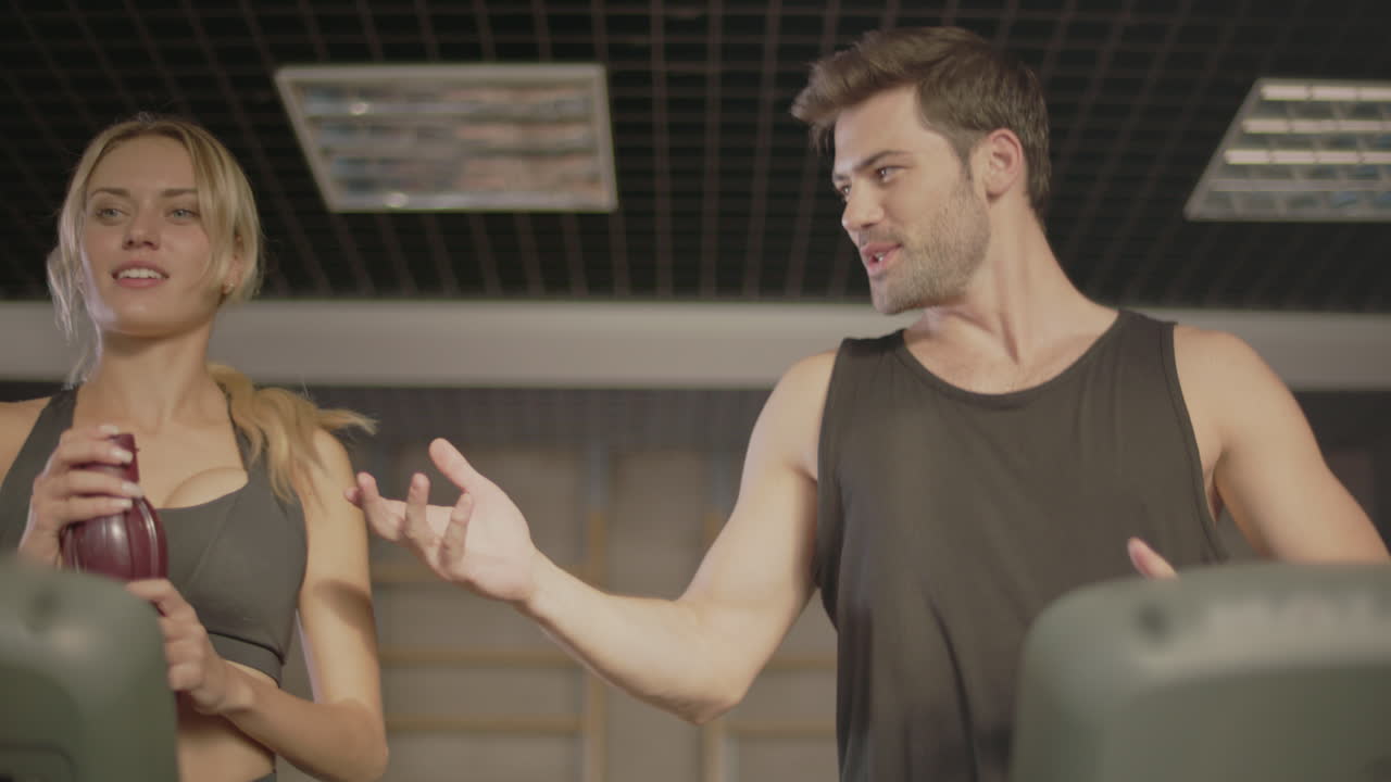 미소 짓는 남성 달리기 선수가 피트니스 체육관에 있는 여자에게 음료 병을 주고 있습니다.