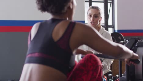 Video-Einer-Vielfältigen-Trainerin-Mit-Tablet,-Die-Eine-Frau-Auf-Einem-Rudergerät-Beim-Training-Im-Fitnessstudio-Ermutigt