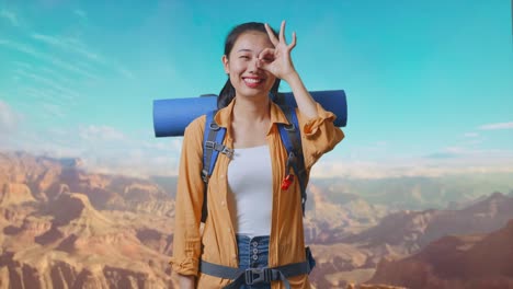 asian female hiker with mountaineering backpack smiling and showing ok sign with fingers over eyes while traveling at the top of mountain