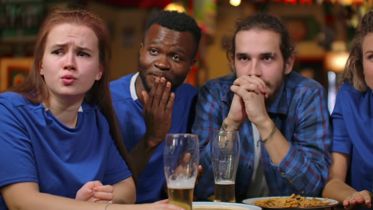 Close-up group of fans emotionally watching a sports broadcast in a bar on TV with beer men and women in blue T-shirts rejoice and hug celebrating a goal scored by their football soccer team.