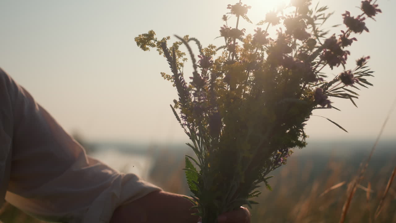 woman squatting in tall grass holding bouquet of wildflowers under golden sunlight with eyes closed and peaceful smile while admiring and scenting blossoms during calm summer