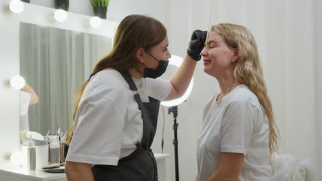 beautician in white top and apron gently picks client eyebrow while both share a smile. wax stick is visible on client forehead, creating light, professional moment inside bright, modern beauty salon