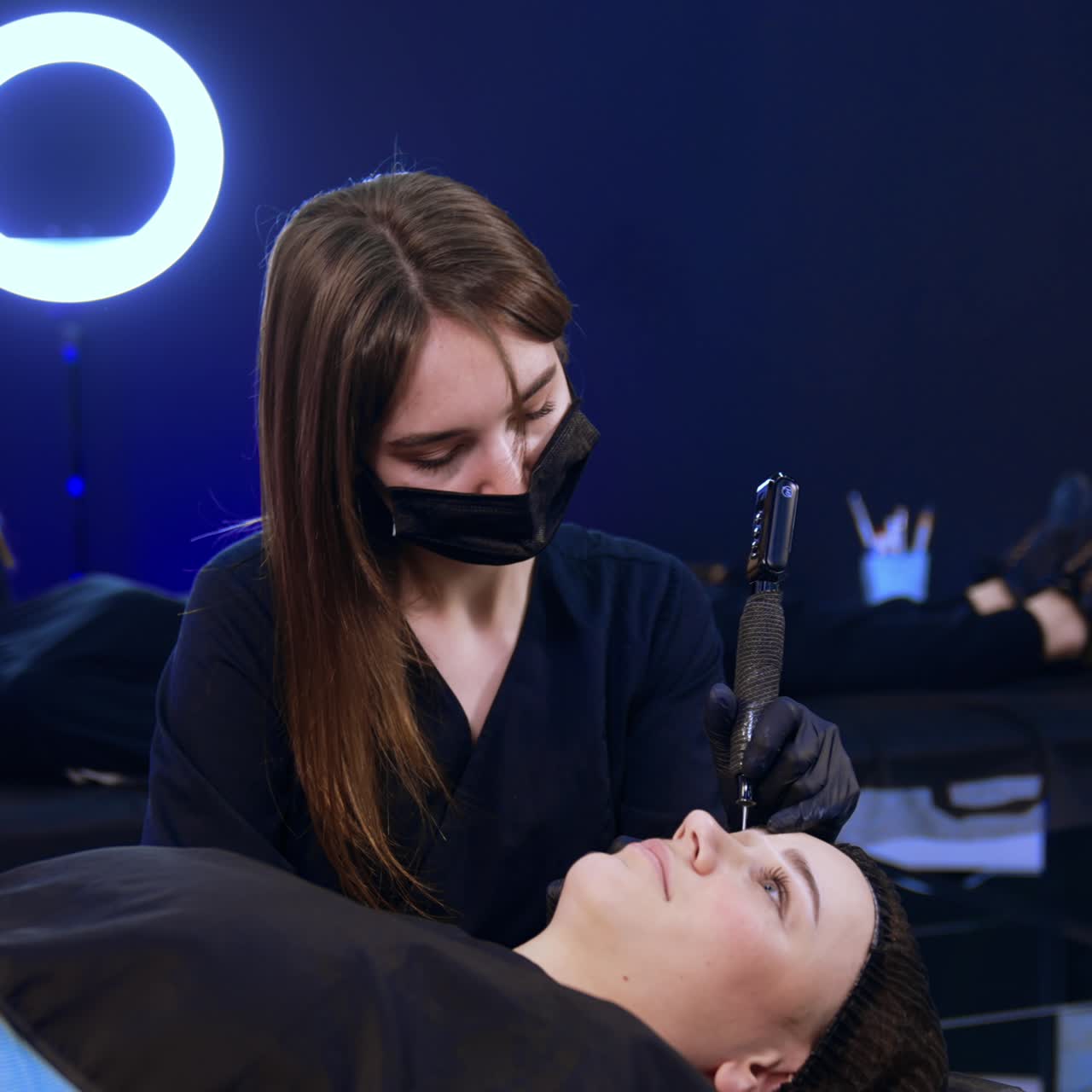 Young female lies on the couch while beautician performs eyebrow procedure for her. Cosmetician uses special device to apply permanent eyebrow make up