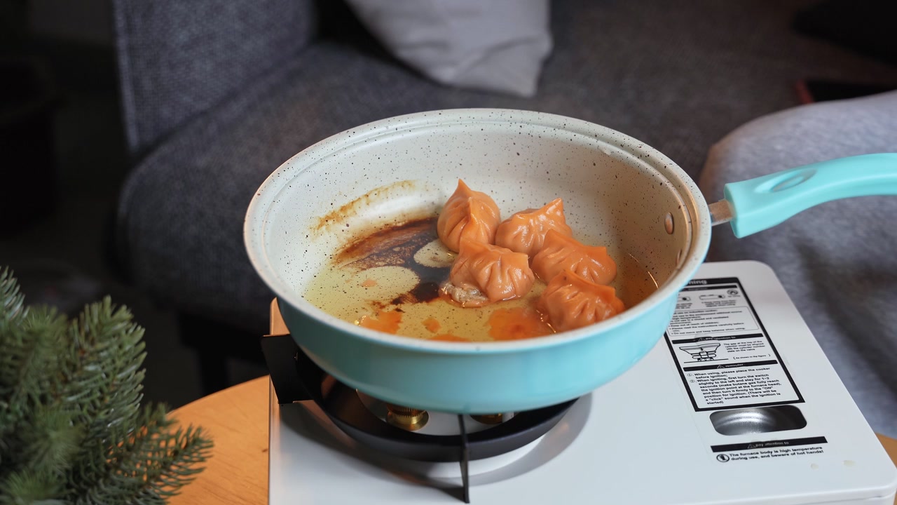 青いフライパンでオレンジ色の餃子を青いヘラと銀色のフォークでひっくり返している手元のクローズアップ。熱いフライパンは、屋内で昼食の準備をしている最中、木製のテーブルに置かれたポータブルガスコンロの上にある。