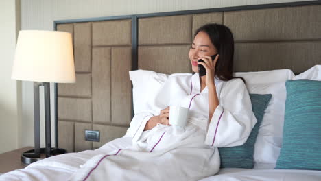 wide shot of young adult asian woman in white bathrobe on a hotel double bed holding a cup of tea coffee and having a joyful happy conversation on her mobile phone