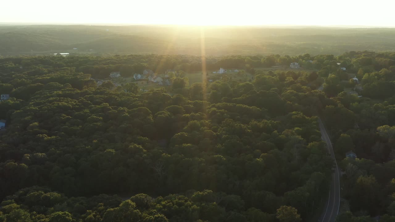 aerial flight towards the bright sunset over a forested rural area of the Northeastern USA, with intense lens flare