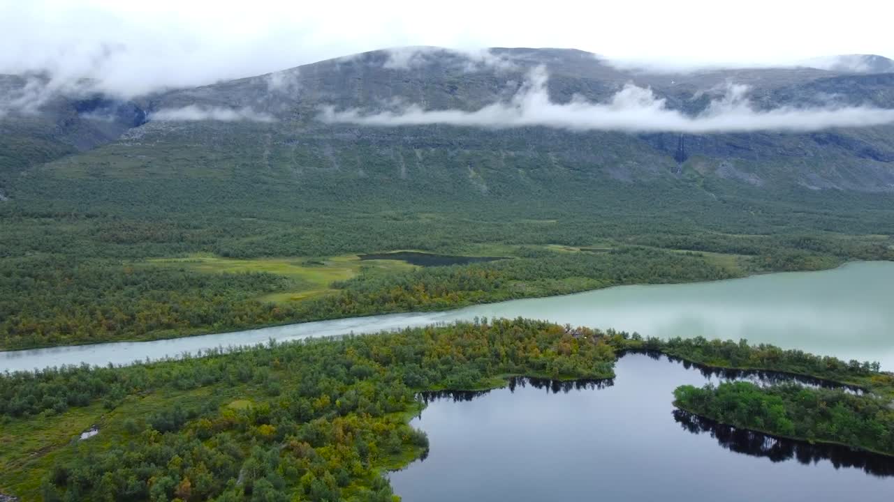 Aerial drone footage of beautiful Sweden bogs or marshlands in front of large mountains during a cloudy day. The lake and rivers and reflective and green trees and nature surround them, white clouds.