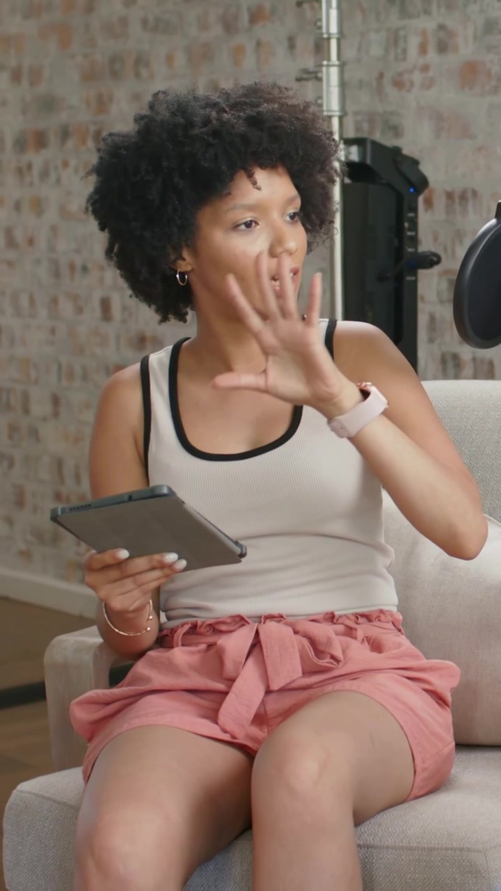 Video verticale: Donna in canotta e pantaloncini rosa che si sposta, seduta sul divano con un tablet, registra con un microfono.