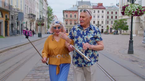 Un-Viejo-Turista-Mayor-Con-Una-Mujer-Caminando-Por-La-Ciudad-Con-Un-Teléfono-Inteligente-En-Un-Palo-Selfie-Teniendo-Una-Videollamada