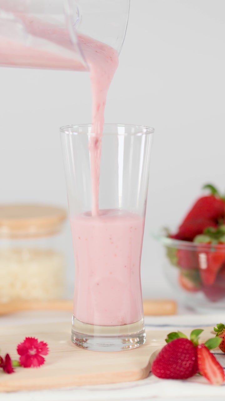 Un frappè rosa alla fragola si versa dolcemente in un bicchiere, circondato da fragole fresche, con luce diurna brillante, inquadratura statica.