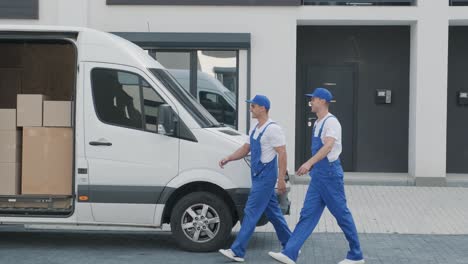 two young workers of removal company unload boxes and furniture from minibus into customer's home