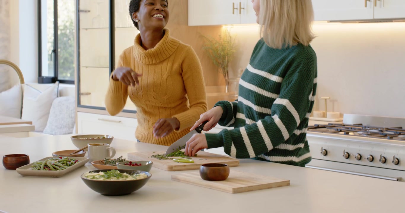 Diverse women in sweaters chopping greens at kitchen island, serving bowls as turkey being set down