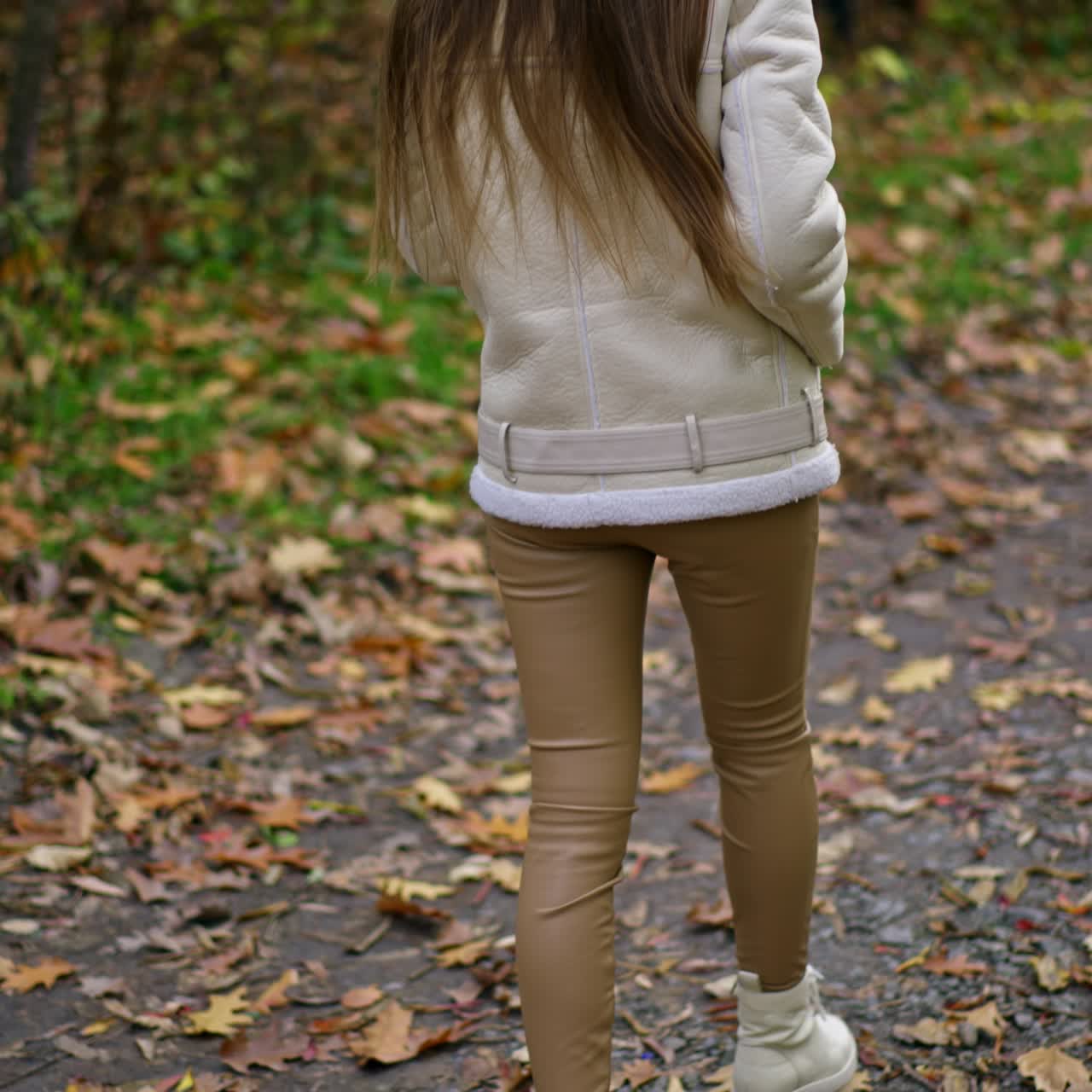 Unrecognized lady in beige pants and white jacket and boots walking in the park. Long-haired brunette turning around demonstrating her look