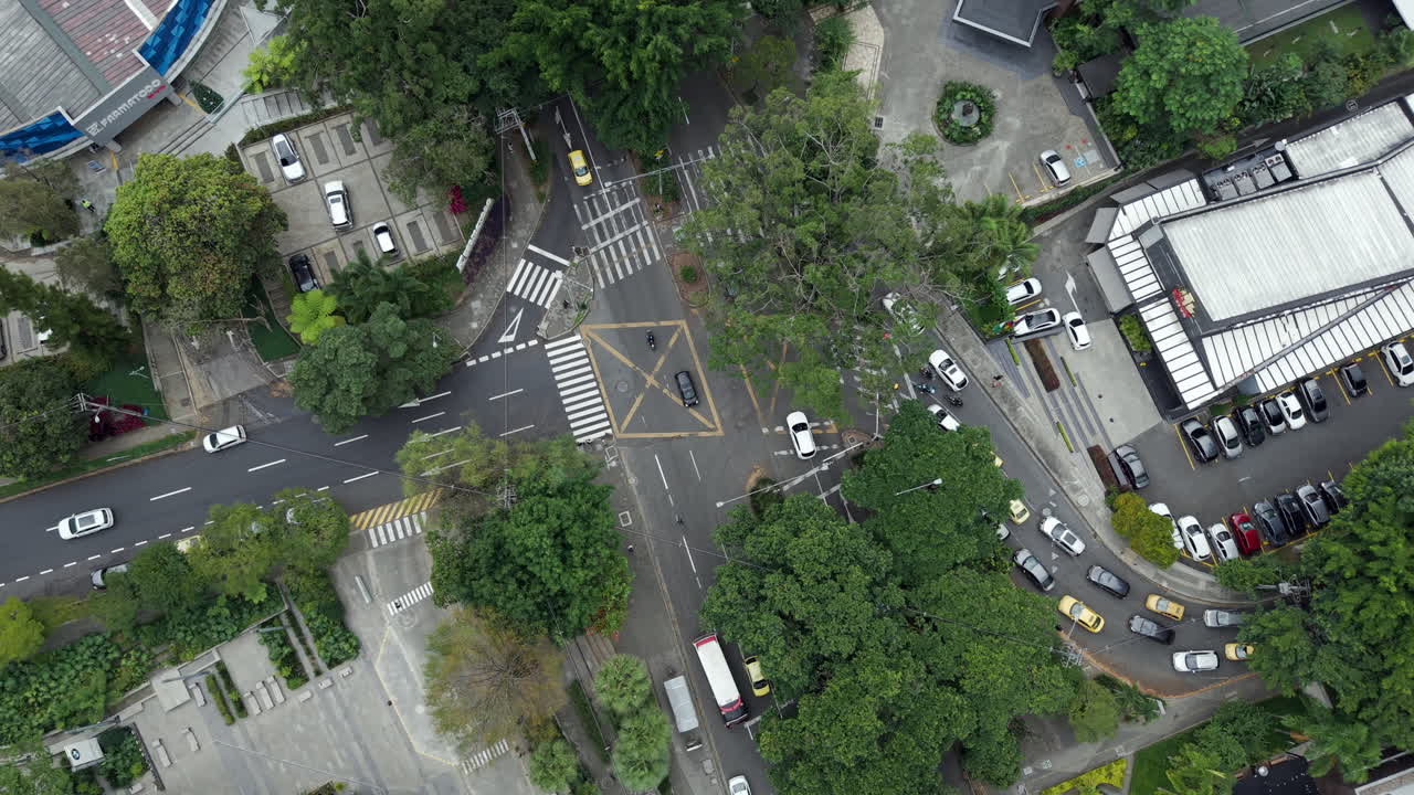 Aerial footage of a bustling intersection in El Poblado, Medellin. Captures the vibrant urban scene with cars navigating through streets surrounded by lush greenery and architecture