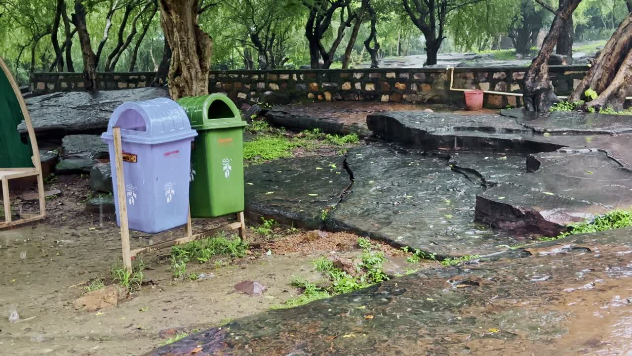 A static shot shows purple and green waste bins standing on rain-soaked ground, surrounded by wet stones, trees, and fresh greenery, reflecting nature’s calm after rainfall