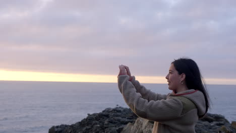 mujer joven grabando el paisaje, una puesta de sol en el océano pacífico, de vacaciones en chile, envuelta en el frío, con una hermosa luz de puesta de sol