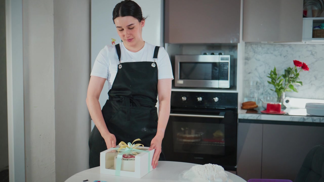 Woman in black apron stands in a modern kitchen. She carefully adjusts a small white box on a table. Her expression is focused and calm. Kitchen background includes oven, microwave