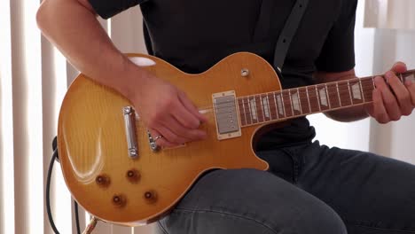 joven en jeans y camisa negra tocando solo de guitarra eléctrica en un taburete contra un fondo brillante