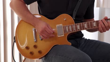 young man in jeans and black shirt playing electro guitar solo on a stool against a bright background