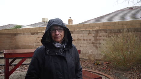 Eine-ältere-Frau-Läuft-In-Einem-Regenmantel-Und-Mit-Kapuze-Während-Eines-Winterregensturms,-Während-In-Zeitlupe-Tropfen-Unter-Einem-Bewölkten-Himmel-Fallen