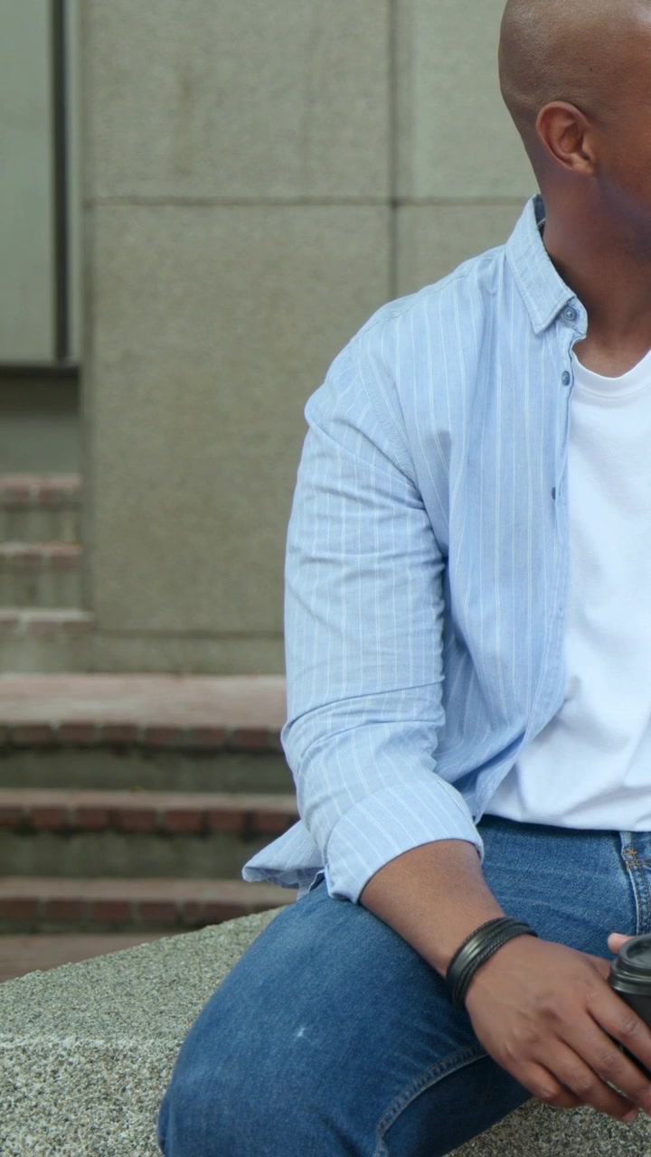 Video verticale: Un uomo in camicia blu lancia un'occhiata, tenendo una tazza da asporto su un davanzale mentre i pedoni passano dietro.