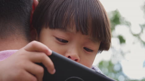 Primer-Plano-De-Un-Padre-Asiático-Abrazando-A-Su-Hijo-En-El-Jardín-Mientras-El-Niño-Mira-Por-Encima-Del-Hombro-El-Teléfono-Móvil