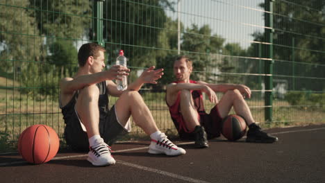 Two-Male-Basketball-Players-Chatting-While-Sitting-And-Leaning-Against-A-Metal-Fence-In-An-Outdoor-Basketball-Court