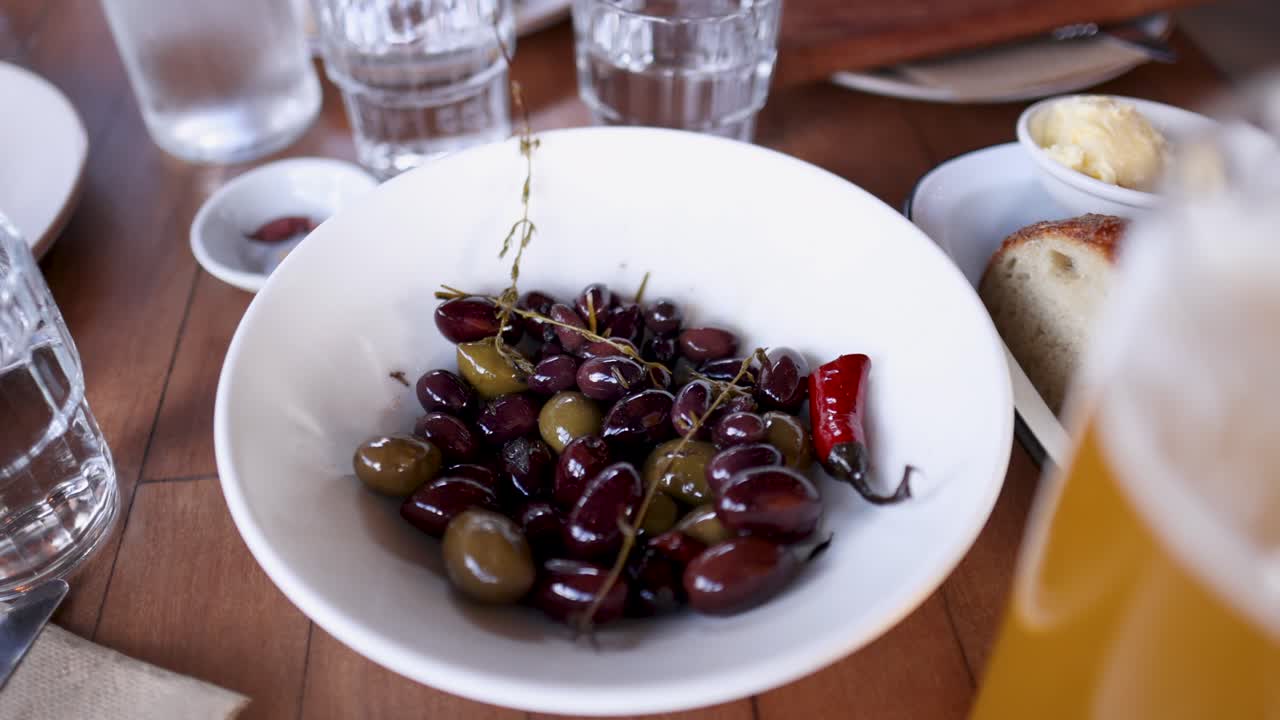 A bowl of marinated olives sits on a wooden table surrounded by drinks and bread in Byron Bay
