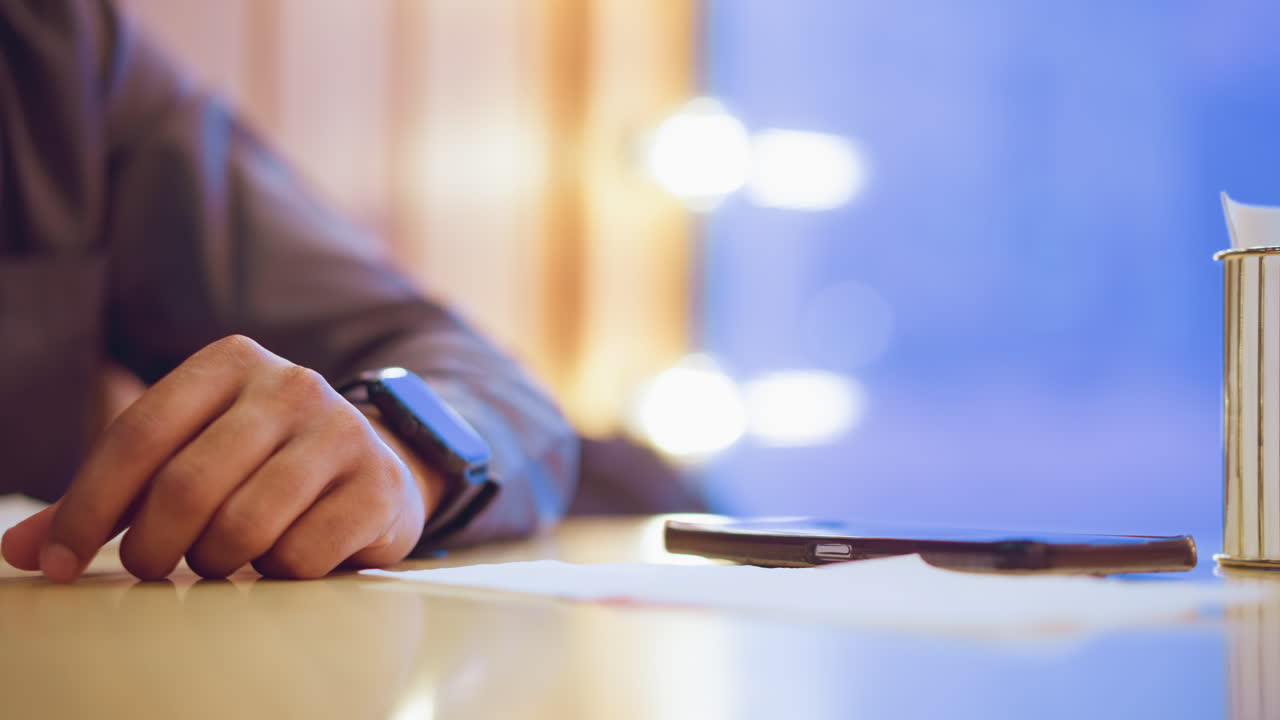 Closeup of person s hand wearing smartwatch resting on table next to smartphone and napkin holder with tissue stack, with blurred evening lights and soft blue background adding cozy ambient atmosphere
