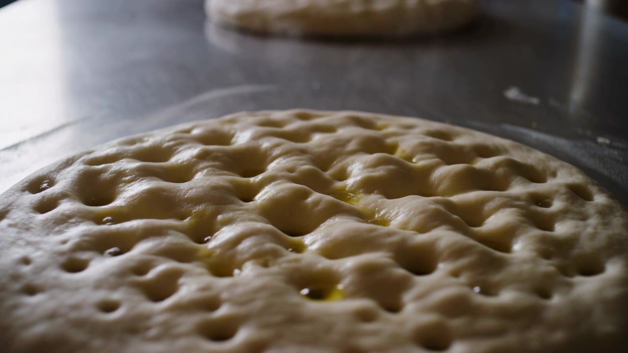 Un primo piano dal basso mostra un impasto di pane lievitato e bucherellato che riposa su una superficie di lavoro in metallo scuro, con fossette create dalle dita uniformemente distanziate, una lucentezza umida e brillante, una leggera spolverata di farina, una forma arrotondata e un secondo impasto sfocato.