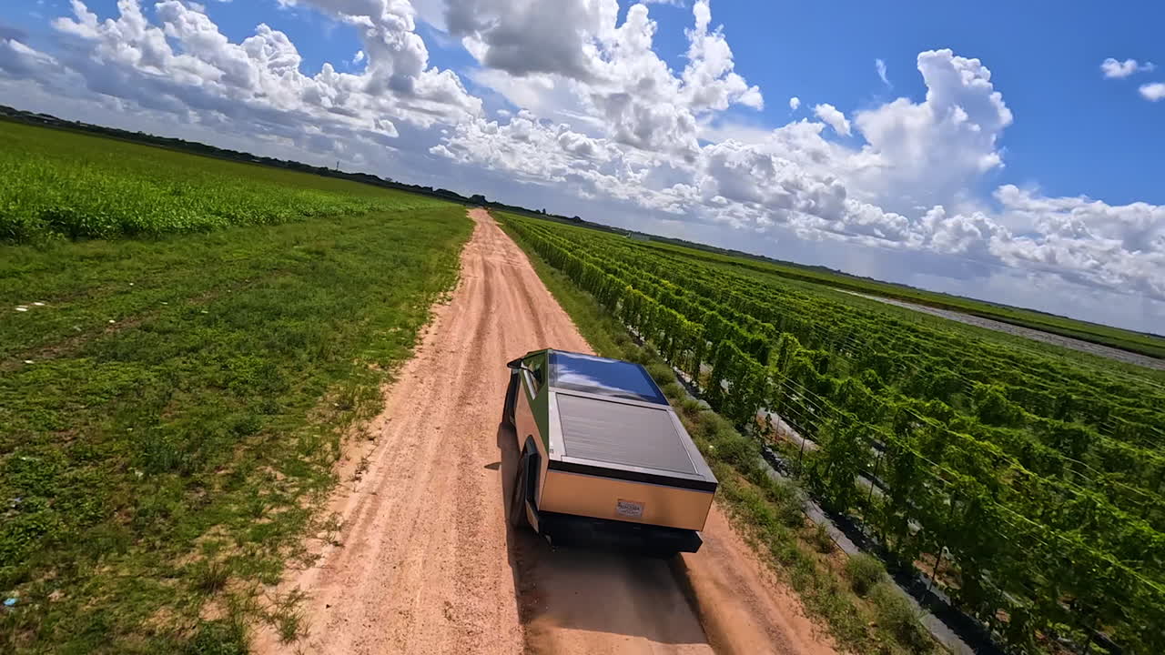 Catching up with the electric car driving fast by the ground road in the farmlands. Aerial perspective on the moving Tesla Cybertruck from FPV drone.