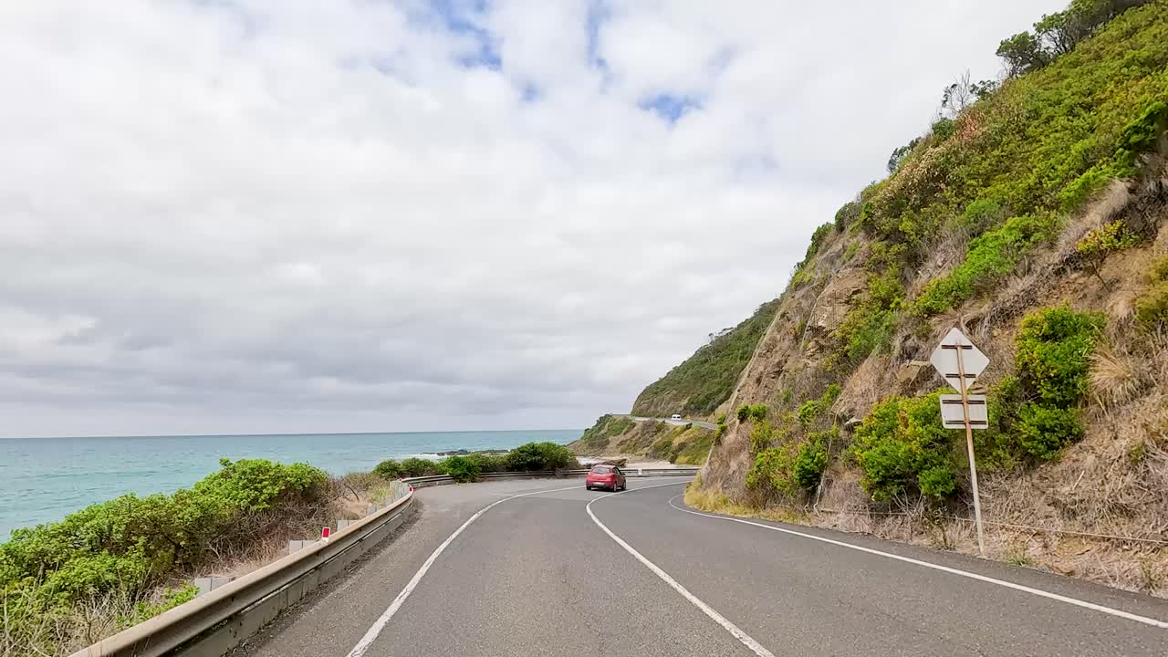 A car travels along the picturesque Great Ocean Road, flanked by lush greenery and ocean views under a cloudy sky