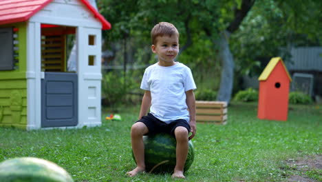 lovely caucasian baby boy sitting on a big water-melon lying on the grass in the yard. cute kid stands up and turns to the berry. playhouse at backdrop.