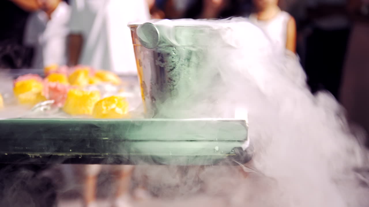 White thick steam goes from the bucket during preparation of ice cream outdoors. People watch the show of making ice-cream with liquid nitrogen.