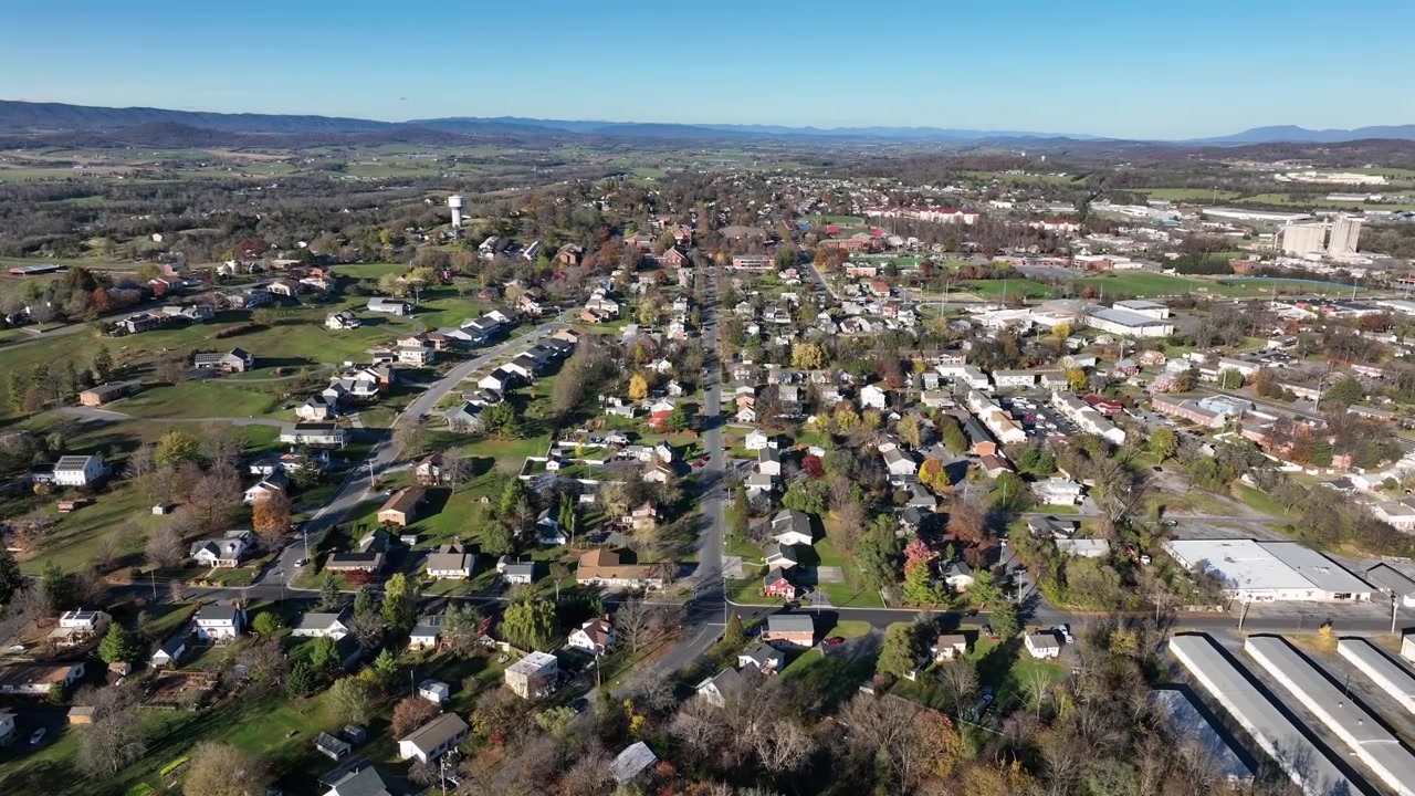 Ripresa con drone in atterraggio di un tranquillo quartiere americano in periferia. Incrocio con strada dritta e case tra alberi colorati. Giornata di sole in autunno. Inquadratura ampia. Colline sullo sfondo.