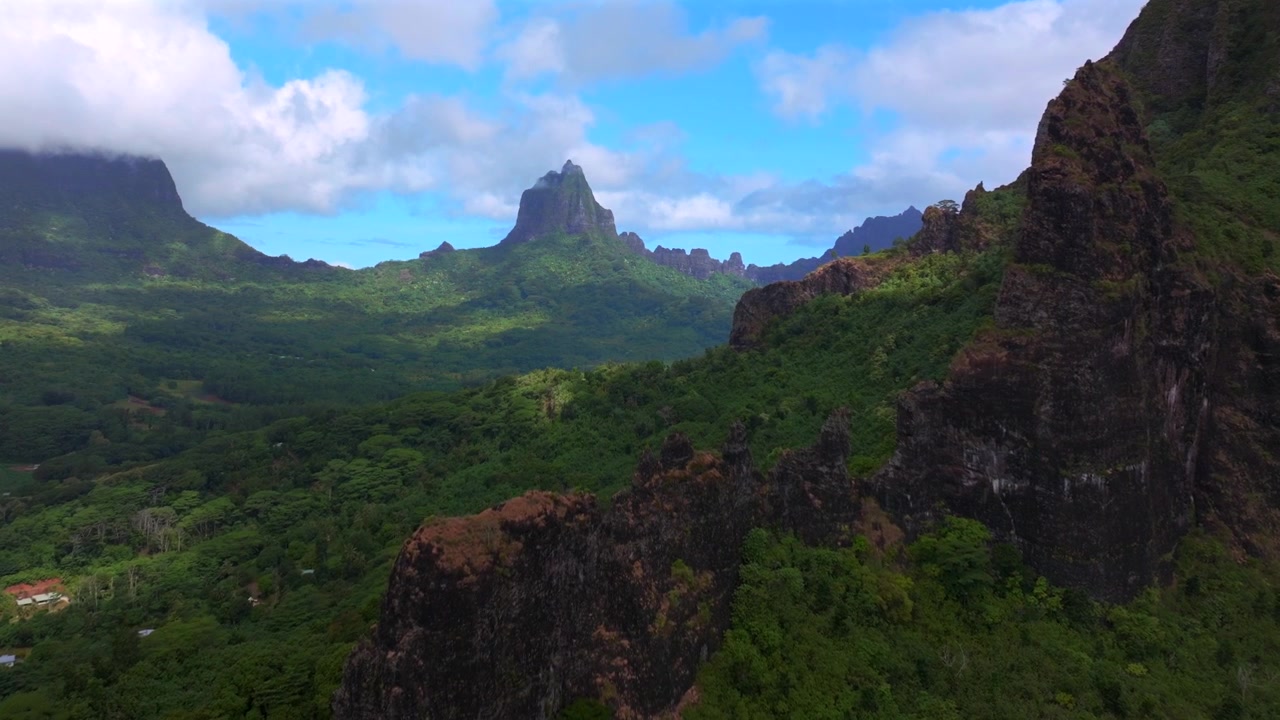 Utsiktsplatsen Belvedere, Moorea, Tahiti, Franska Polynesien, berget Toheia, berget Rotui, flygfoto med drönare, frodig grön djungelskog, vulkaniska, taggiga, oländiga bergstoppar, solig morgon, blå himmel, moln, panorerar framåt och uppåt.