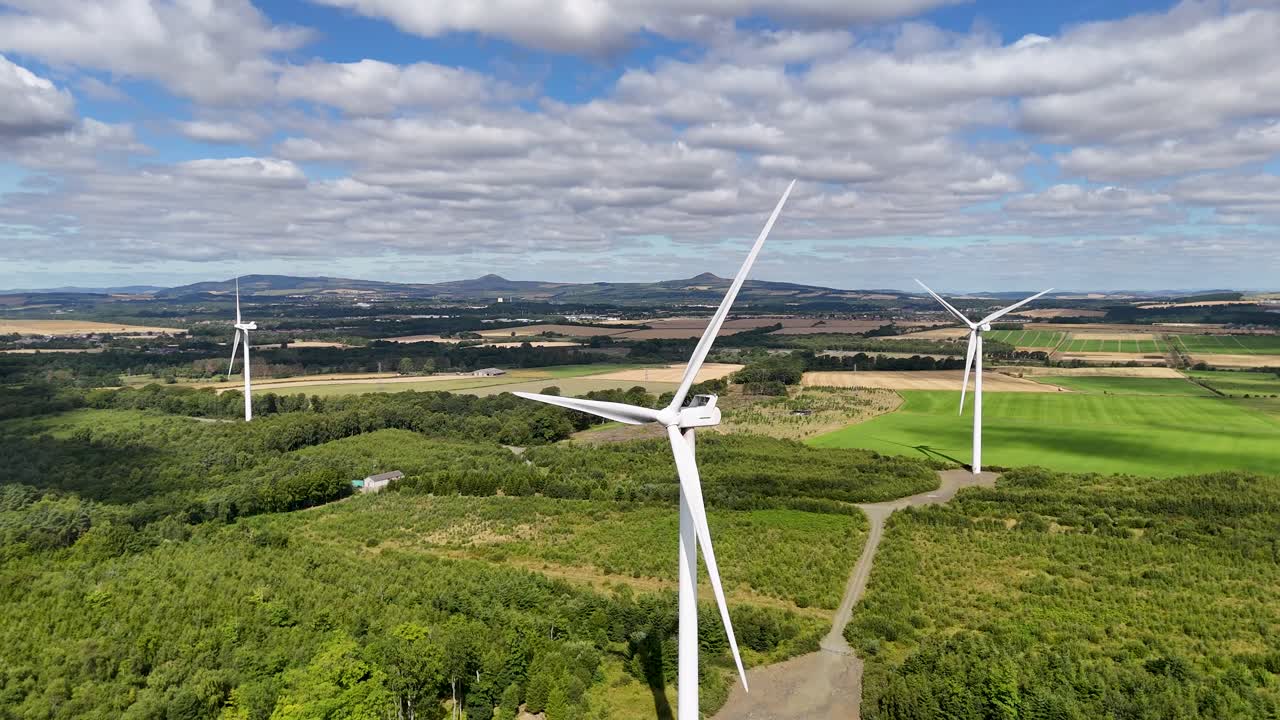 Drone footage captures wind turbines rotating above green fields and forests in Fife, Scotland, under bright daylight with partly cloudy skies