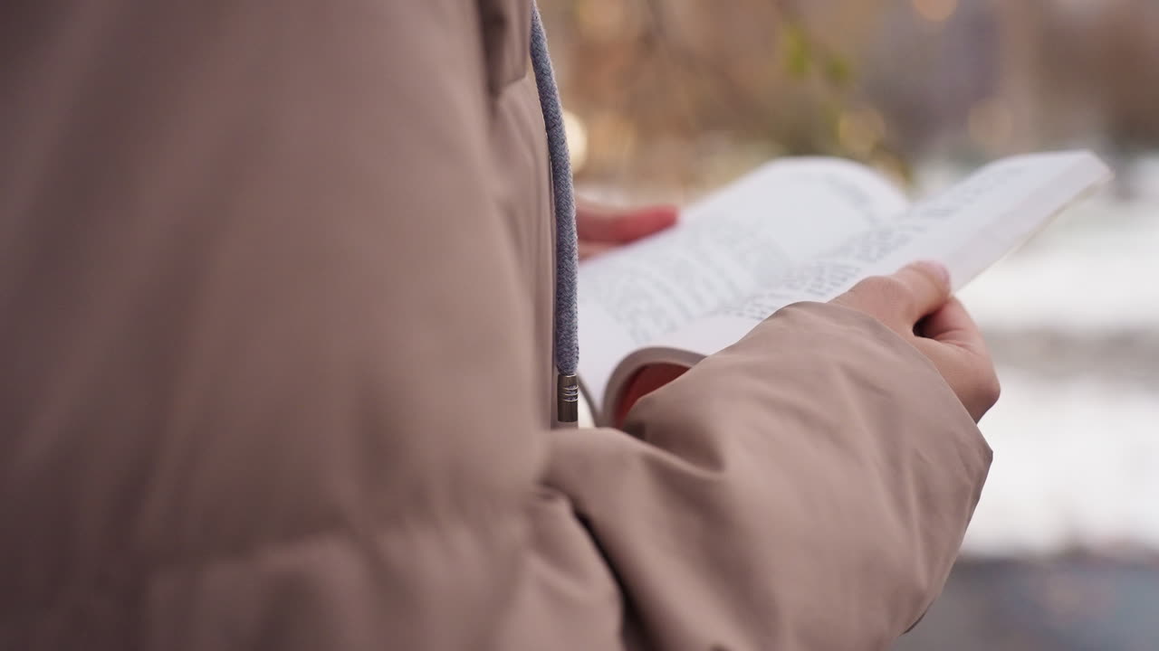 Side view of person wearing winter sweater holding open book outdoors during cold weather, snow-covered landscape and trees in background create peaceful atmosphere with soft light